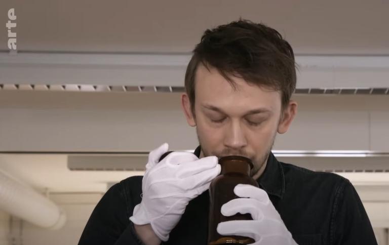 Ein Mann riecht an einer Flasche, welche den Geruch einer Frau auf einem Wattebausch enthält.