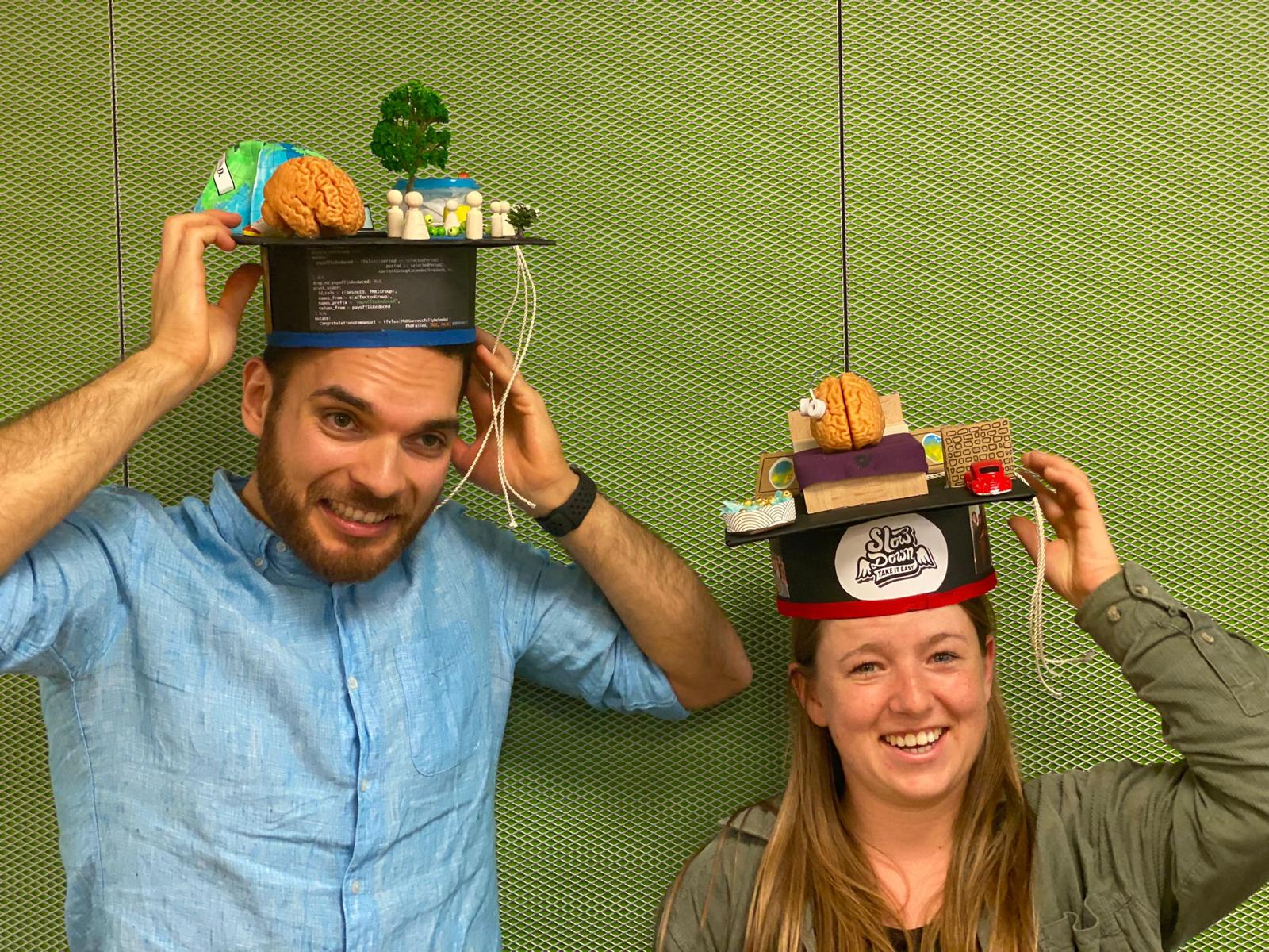Emmanuel and Mirjam wear their doctoral hats, which represent the contents of their dissertations, e.g. a bed and brain on Mirjam's hat and a brain and globe on Emmanuel's hat.