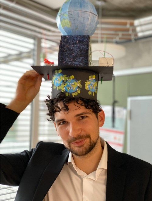 A photo of Benedikt Langenbach wearing a doctoral hat, which shows important aspects of his dissertation, e.g. a picture of his brain, a blue planet, and tDCS electrodes.